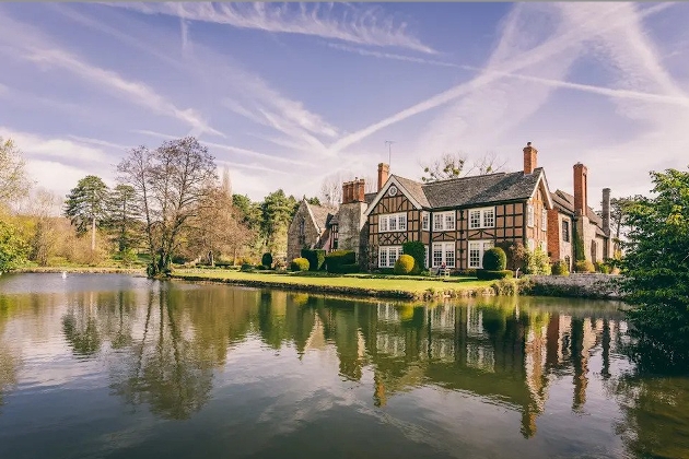 The exterior of a large manor house surrounded by gardens and a lake